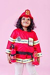 Child wearing a red firefighter costume and helmet stands on a pink background holding toy fire-safety items.