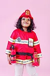 Child wearing a red firefighter costume and helmet stands on a pink background holding toy fire-safety items.