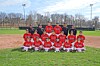 A youth baseball team poses in red uniforms on a baseball field with coaches standing behind them.