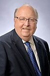 Studio portrait of a man wearing glasses, a dark suit and blue tie on a light background.