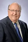 Studio portrait of a man wearing glasses, a dark suit and blue tie on a light background.