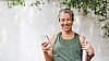 Smiling woman holding a smartphone outdoors beside a white wall and hanging green branches.