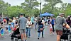 Crowd of people, including children, gathered outdoors near a blue canopy tent in a park.