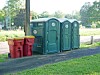 Portable toilets next to red trash bins along a paved path in a grassy park area.