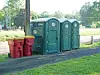Portable toilets next to red trash bins along a paved path in a grassy park area.
