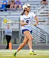 A lacrosse player in white uniform and helmet runs with a lacrosse stick on a field.