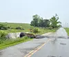 Floodwater crosses a rural road bordered by grass and trees on an overcast day.