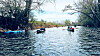 Several rowboats with people on a tree-lined river under a bright sky.