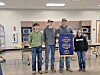 Four students stand in a cafeteria holding a blue banner, with tables and cabinets behind them.