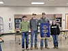 Four students stand in a cafeteria holding a blue banner, with tables and cabinets behind them.