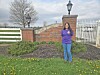 Woman standing on grass near a brick wall, white fence, and landscaped plants outdoors.