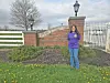Woman standing on grass near a brick wall, white fence, and landscaped plants outdoors.