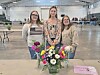 Three women stand behind a vase of colorful flowers in a large indoor hall.