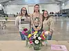 Three women stand behind a vase of colorful flowers in a large indoor hall.