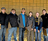 Six people standing together indoors in front of a tan tiled wall.