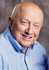 Smiling elderly man in a blue checkered shirt against a gray backdrop.