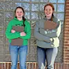 Two women standing outdoors, each holding a clipboard in front of a glass block wall.