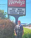 A man stands beside a Wendy's roadside sign with a congratulatory message about a gold medal.