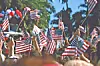 Crowd waving many American flags outdoors with palm trees in the background.