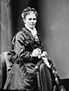 Black-and-white portrait of a Victorian-era woman seated beside a chair in a studio.