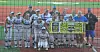 Youth baseball players and coaches pose on a baseball field behind a yellow sign.