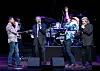 Four musicians sing and play on stage in front of a drum kit and amplifiers.