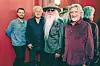 Five men posing indoors against a red backdrop, including one man with a long white beard, hat, and sunglasses.