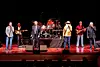 Several musicians perform on a stage with drums, guitars and microphones in front of a dark audience.