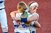 Two athletes in white uniforms embrace on a dirt field with a red wall behind them.