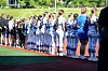 Softball players in uniforms stand lined up along a field near the dugout area.