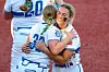 Two softball players in white uniforms embrace on a red dirt softball field.