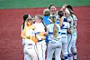 A group of softball players in white uniforms huddles on the field.