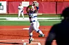 Softball player in white uniform reaches up with a glove to catch a ball near home plate.