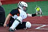 A softball catcher kneels behind home plate and reaches for a ball near the strike zone.