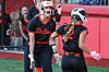 Two softball players in black and orange uniforms standing on a field near a fence.
