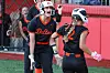 Two softball players in black and orange uniforms standing on a field near a fence.
