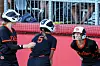 Three softball players in helmets stand near a red wall and chain-link fence at the field.