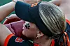 Softball player in a black face guard and orange-trimmed jersey, shown in close-up on the field.
