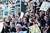 Crowd seated in bleachers holding signs at an outdoor event.