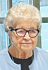 Elderly woman with white hair and black glasses indoors against a blue background.