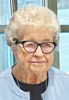 Elderly woman with white hair and black glasses indoors against a blue background.