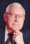 Studio portrait of an elderly man in glasses and a suit, with his hand under his chin.