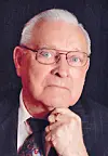 Studio portrait of an elderly man in glasses and a suit, with his hand under his chin.