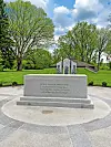 Granite memorial stone in a park with trees, grass, and a small monument in the background.