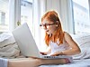 Woman with glasses using a laptop while sitting on a bed by a window.