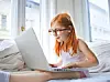 Woman with glasses using a laptop while sitting on a bed by a window.