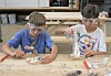 Two children in safety goggles work on small projects at a wooden table in a workshop.