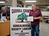 Man holding a certificate next to a Carroll County Genealogical Society Library sign indoors.