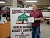 Man holding a certificate next to a Carroll County Genealogical Society Library sign indoors.