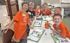 Children in orange shirts gather around a table covered with books, papers, and small orange containers in a classroom.
